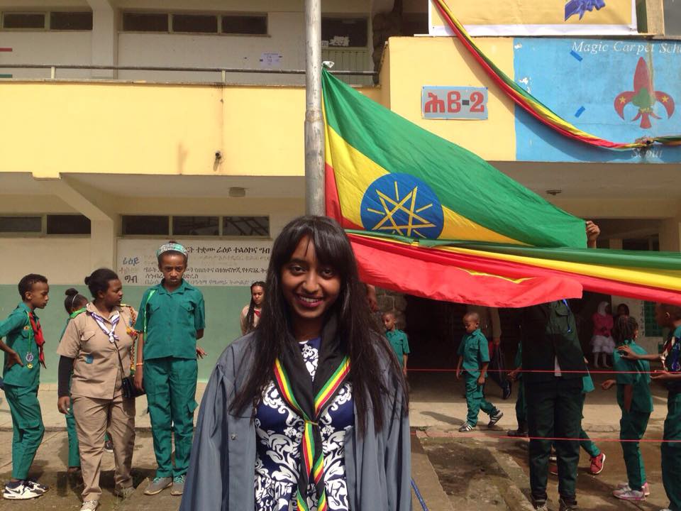 Saron Gebre in front of the Ethiopian flag pole, adorned in her graduation gown and scout scarf. In the background, her fellow scout sisters and brothers join her in the momentous occasion.